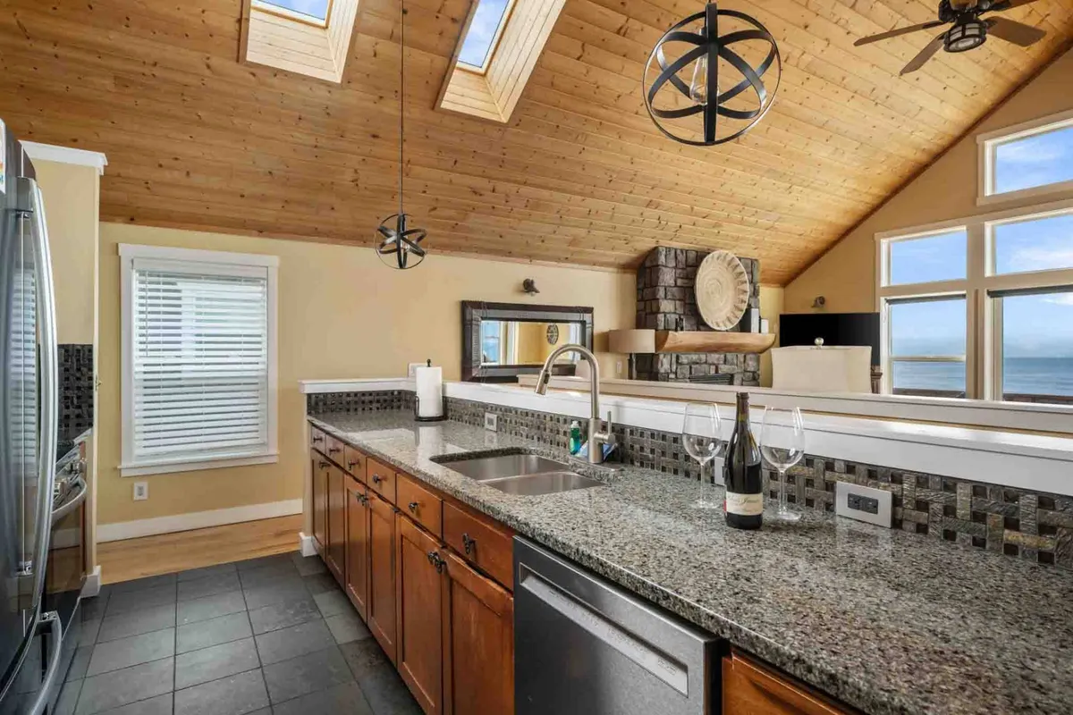 A spacious kitchen and dining area with a granite countertop, large windows bathing the space in natural light, elegant lighting fixtures, and a wooden ceiling. The open layout connects the kitchen to the dining area, offering views and a setting ideal for both casual breakfasts and sunset dinners.