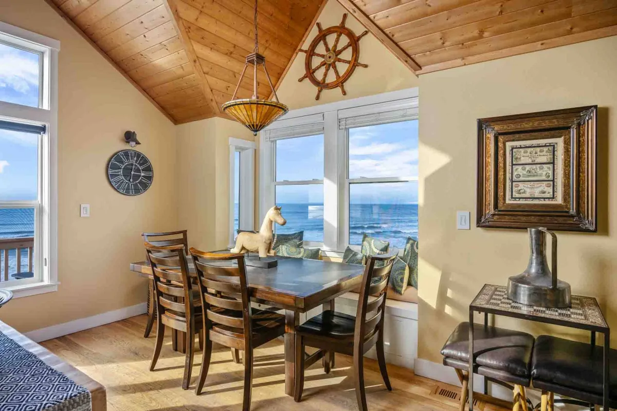 A cozy dining area with a wooden table set beside large windows overlooking the ocean. Nautical décor, including a ship's wheel and playful figurines, infuses the space with coastal character. The setting is perfect for shared meals and heartfelt conversations with friends and family.