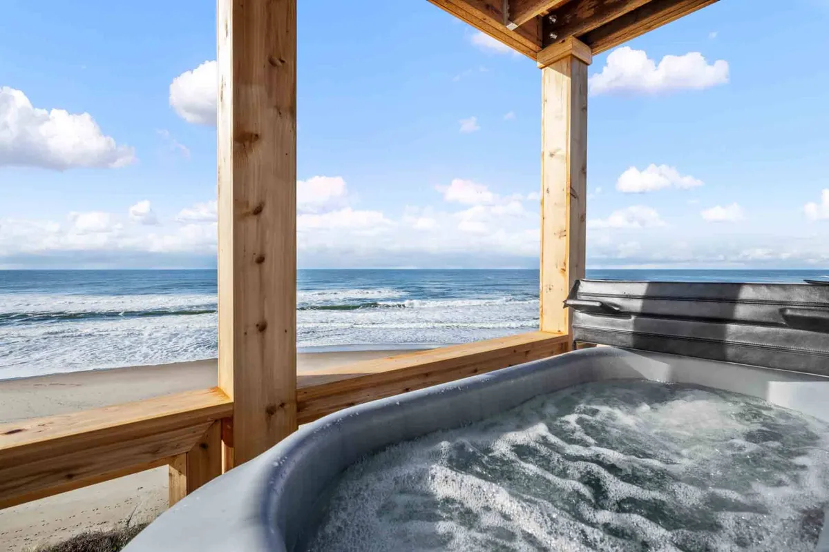 This image features a private hot tub situated on a wooden deck overlooking the vast Pacific Ocean. The hot tub is positioned to maximize ocean views and is complemented by the serene ambience of the surrounding nature, making it an inviting spot for relaxation at any time of day. The scene suggests the ease of moving between the home’s interior, the deck, and the outdoors.