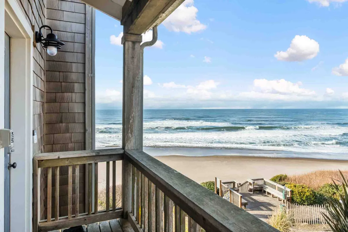 A sweeping view from the balcony showcases the vast ocean, sandy beach, wooden railing, and a dramatic sky dotted with clouds. The picture invites the viewer to imagine themselves relaxing outside, breathing in the fresh ocean air or watching waves no matter the season.