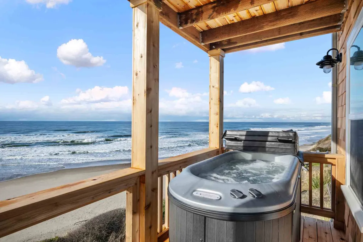 A tranquil shot of the wooden oceanfront deck, with a hot tub overlooking the surf and waves. The deck also features seating, inviting guests to enjoy sunrises, sunsets, or simply breathe in the ocean air. The integration of the hot tub into the deck design underscores the seamless blend between indoor comfort and outdoor adventure.
