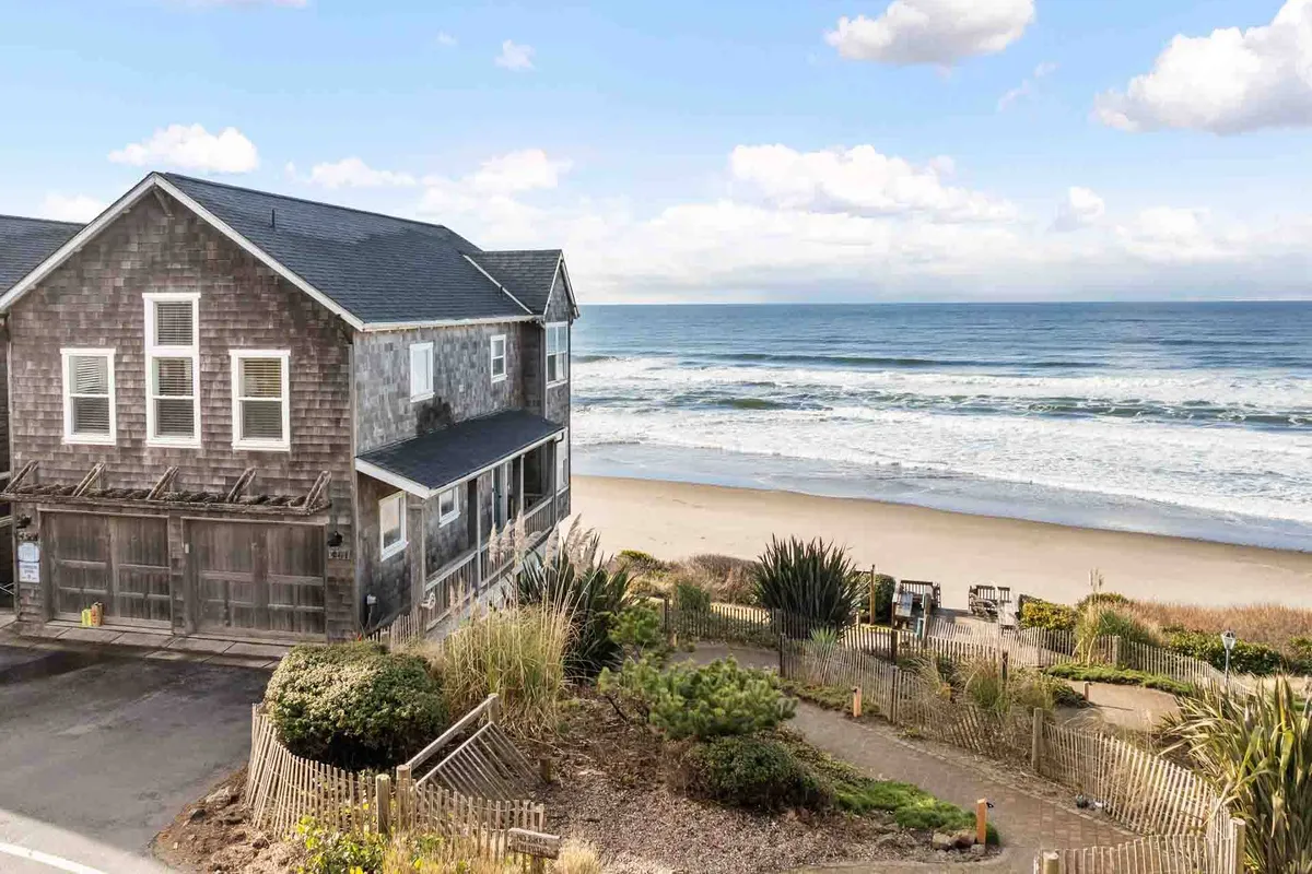 A wide shot of the exterior of Into the Mystic, set against the backdrop of the beach and ocean. The home's welcoming facade is surrounded by a well-kept garden, with sandy paths leading directly to the beach mere steps away.