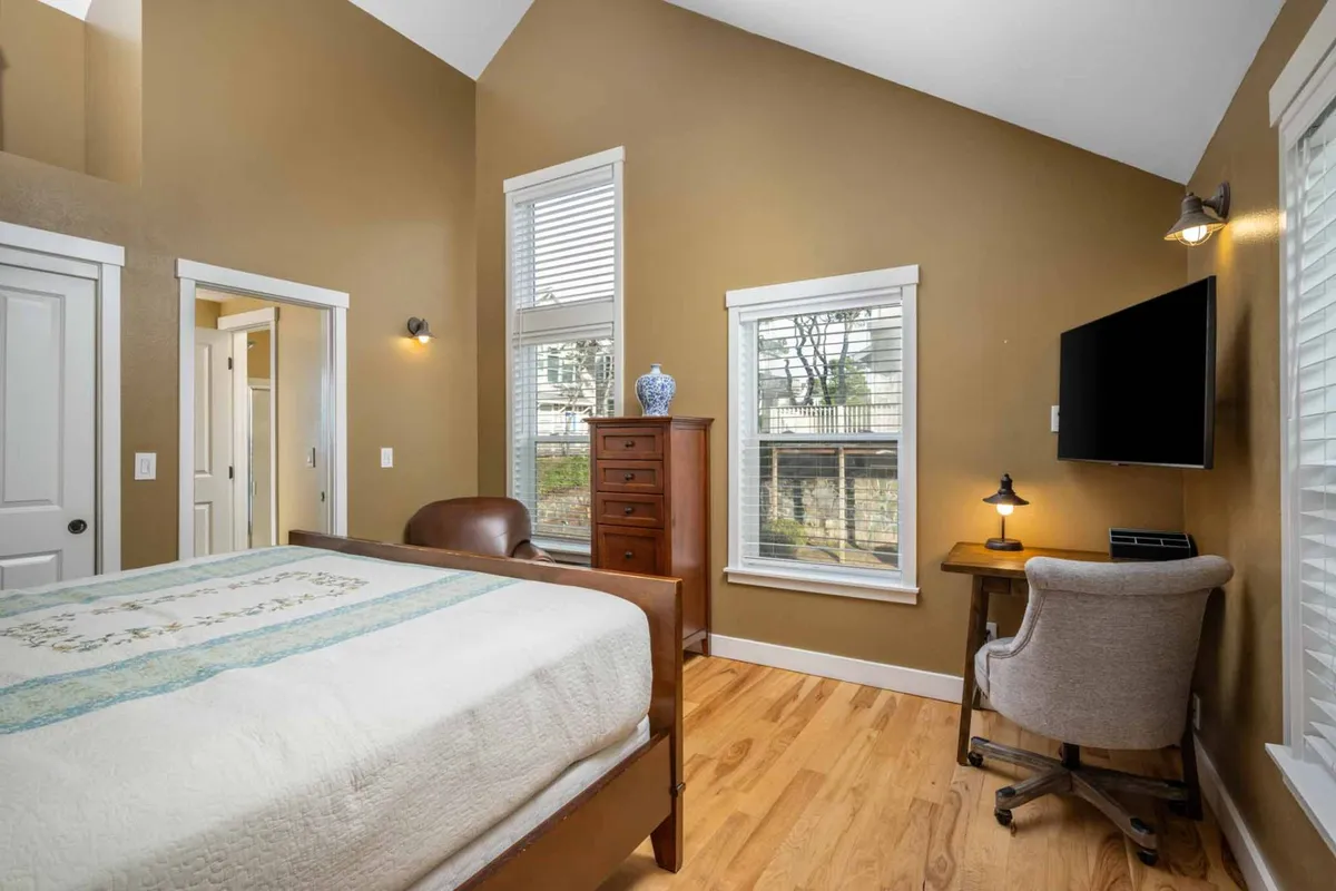 A serene bedroom space featuring a bed, a wooden dresser, a desk and chair by a window, and gentle natural light filtering in. The scene is ideal for relaxation, reading, or remote work in a peaceful environment.