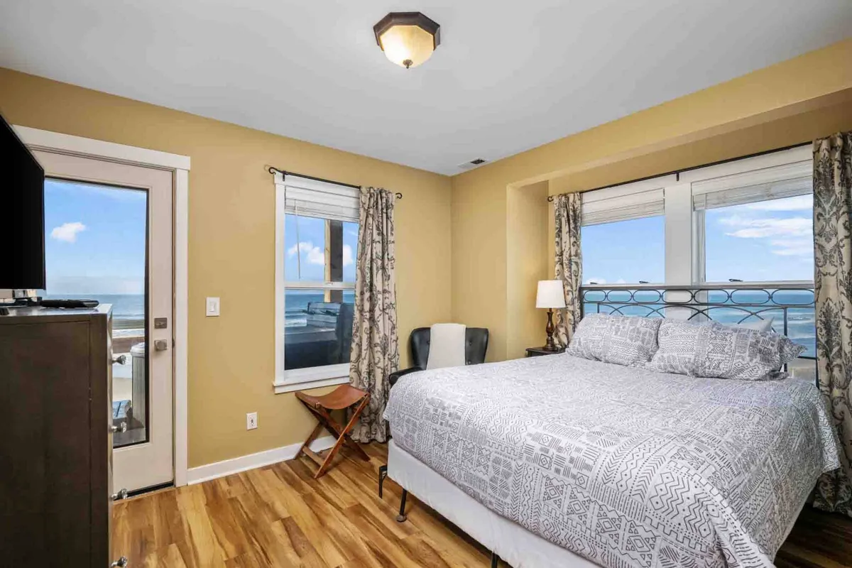 A tranquil bedroom with a bed, windows offering ocean views, decorative curtains, a nightstand with a lamp, and a wall-mounted TV. The space is light-filled and perfect for unwinding.