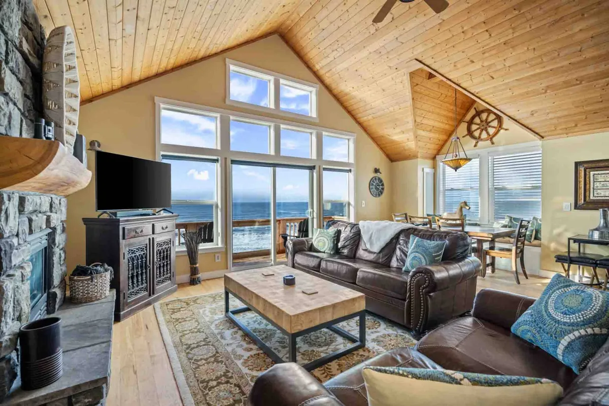 This image showcases a spacious living room with large windows offering sweeping ocean views. The room features a stone fireplace, wooden ceiling, and comfortable leather sofas arranged around a coffee table, creating a cozy and inviting atmosphere. Natural light floods the space, highlighting the coastal décor and making it ideal for gatherings and relaxation.