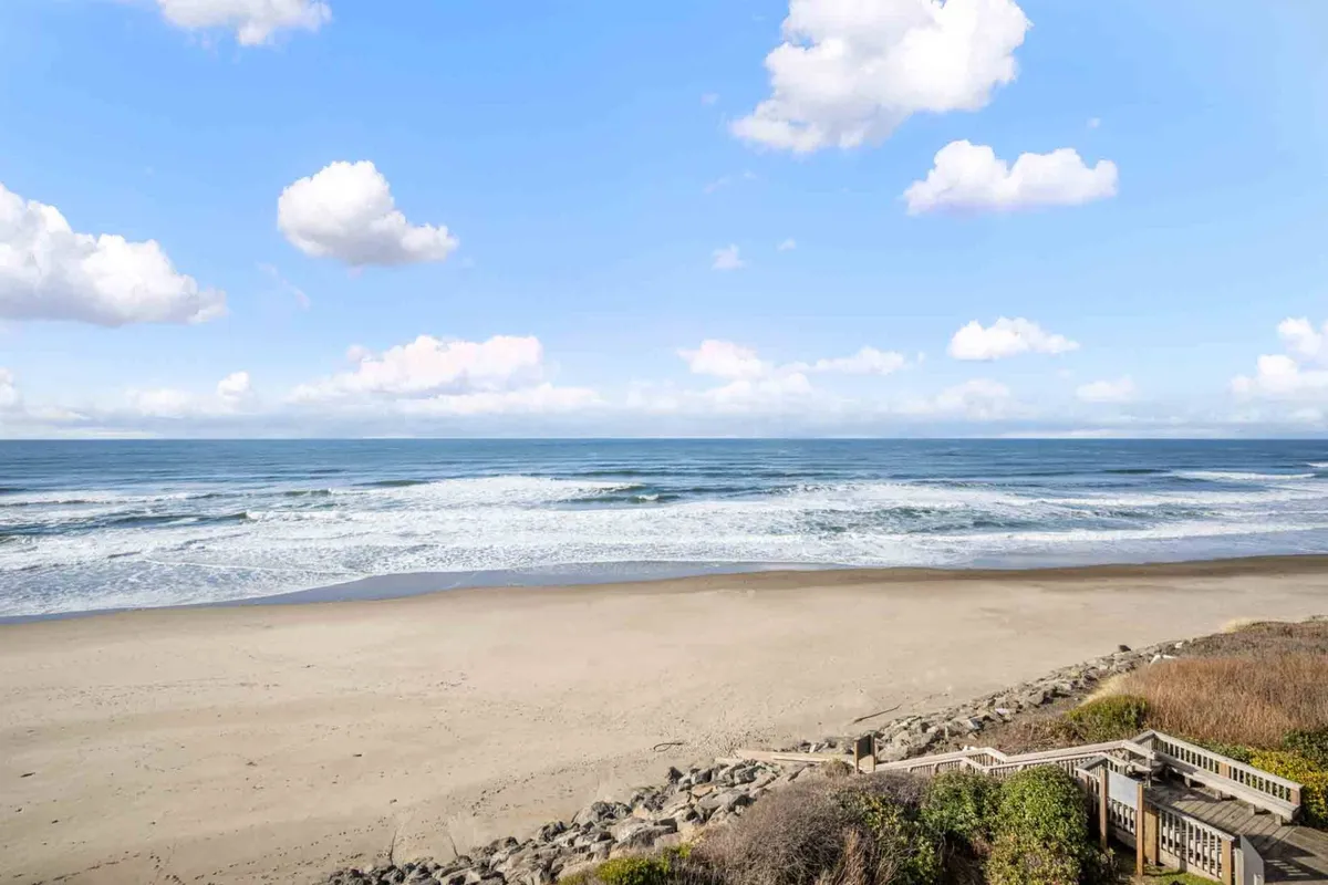 A wide view of the Oregon coastline featuring gentle ocean waves rolling onto a sandy beach, with soft clouds stretching across the sky. The beach is expansive and inviting, perfect for long walks, beachcombing, or simply soaking up the tranquil seaside atmosphere.