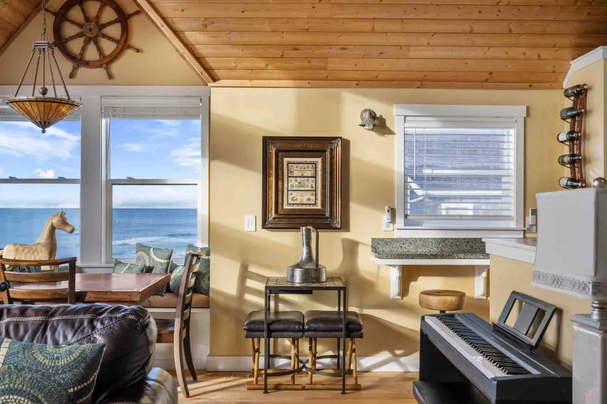 A bright, open interior with expansive windows framing the ocean beyond. The space includes a wooden ceiling, decorative wall art, and a visible piano, emphasizing the musical moments mentioned in the section. The arrangement suggests both a comfortable living and dining area, with thoughtful coastal touches enhancing the ambiance.