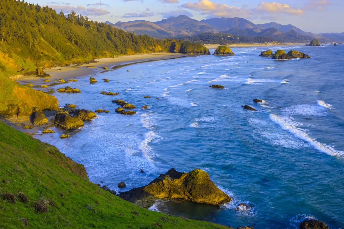 A sweeping coastal view featuring waves crashing on rugged rocks, a sandy beach, lush green cliffs, and distant mountains. The vibrant landscape offers a sense of space to explore and unwind, reinforcing the area's natural beauty.
