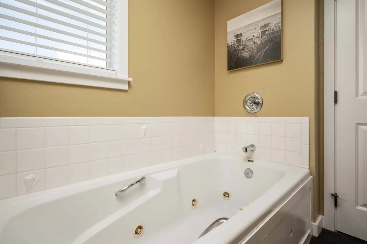 A sunlit bathroom featuring a large whirlpool tub beneath a window with blinds, framed wall art, and soft, serene lighting. The clean lines and spa-like setup evoke luxury and relaxation.