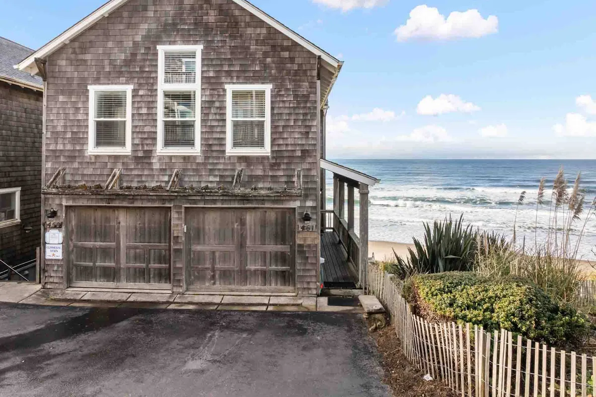 This image presents the weathered wooden exterior of the home, featuring garage doors, touches of surrounding greenery, and a glance at the ocean beyond. The scene radiates tranquility and a strong sense of place at the water's edge, effortlessly capturing the home's oceanfront positioning and connection to the outdoors.