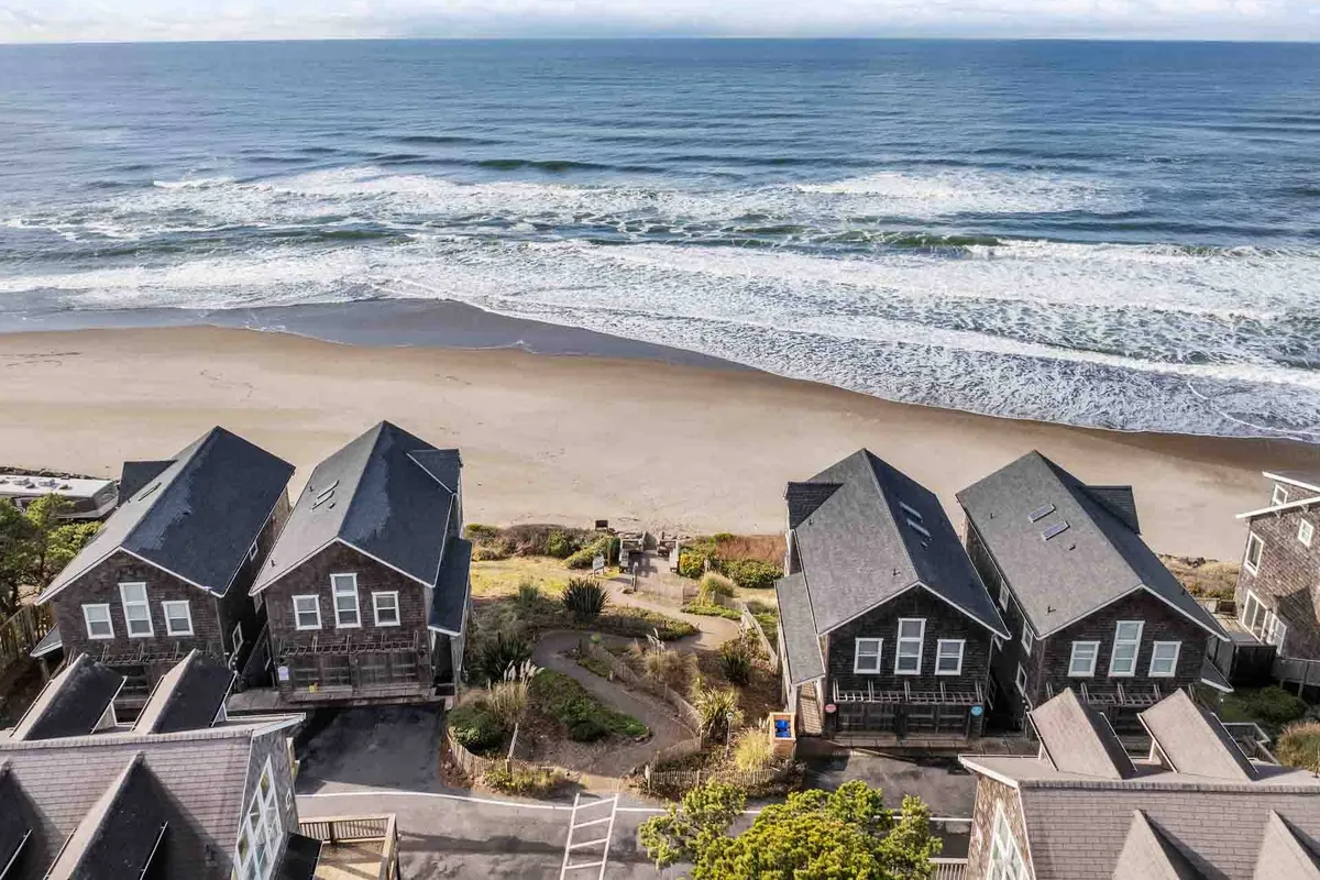 A scenic view of the coastline with gentle ocean waves, a sandy beach, and a pathway leading past several coastal houses. The tranquil seaside setting and open space invite walking, playing, and relaxing outdoors.