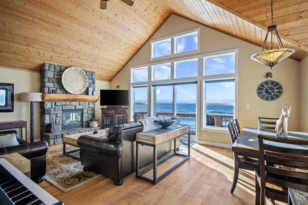 The image showcases a stunning living and dining area with expansive windows revealing breathtaking ocean views. A grand stone fireplace anchors the cozy space, complemented by a wooden ceiling that adds warmth and character. The dining table invites intimate meals, while a plush sofa set offers plenty of space to relax and connect. The overall mood is welcoming and serene, epitomizing the ideal coastal retreat.