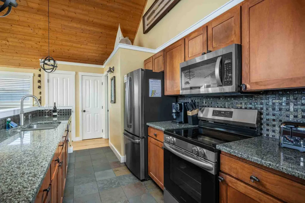 A bright, inviting kitchen featuring a granite countertop, stainless steel appliances, and a rustic wooden ceiling. The open space is designed for group cooking and entertaining, with ample room for meal preparation and gathering around the kitchen table.