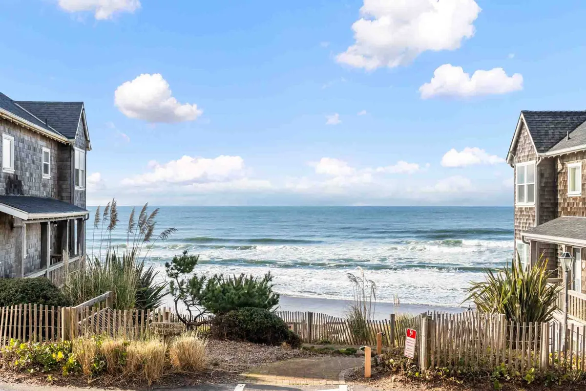This view features the expansive beachfront, rolling ocean waves, nearby homes, a fence, and lush vegetation in the foreground. It evokes the spirit of beach walks, sunbathing, and sunset picnics, all within easy reach of the property.