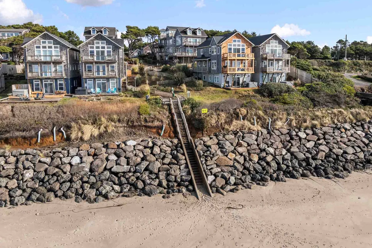 Beachfront houses perched above a wide sandy stretch, with a staircase leading directly down to the beach. The foreground shows the rocky shoreline, while the background includes sunlit houses and the vast coast, suggesting easy access and a family-friendly environment.